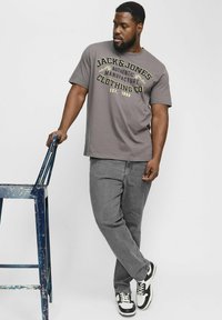 Gray t-shirt featuring black and yellow text, paired with gray jeans and black-and-white sneakers. Subject leans on a blue stool.