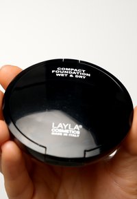 Svart kompakt foundation-fodral med en glänsande yta. Text på locket läser "KOMPAKT FOUNDATION FÖR VÅT OCH TORR ANVÄNDNING" och "LAYLA COSMETICS MADE IN ITALY."
