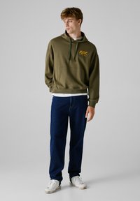 Haut à capuche vert olive avec poche avant et cordon de serrage, arborant un logo jaune. Associé à un jean bleu foncé et des baskets blanches avec des accents verts.