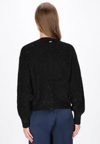 Zwarte ribbeltrui met een losse pasvorm, versierd met lichte glittertjes en een ronde halslijn. De achterkant heeft een eenvoudige, strakke uitstraling.