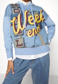 Niebieska bluza z kapturem, ozdobiona graficznym nadrukiem neonowych znaków z napisem "Week end". Zawiera kaptur i ściągacze przy mankietach, sparowana z jasnoniebieskimi dżinsami.