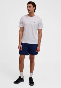 Vit sport-t-shirt med korta ärmar och en liten logga på bröstet, kombinerad med marinblå shorts. Svarta löparskor och vita strumpor.