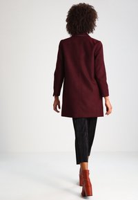 Burgundy ullblandad kappa med rak skärning, långa ärmar och en vent bak, bärs över plisserade svarta byxor och ankelstövlar.