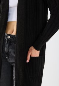 Lange zwarte cardigan met ribbeltextuur, open voorkant en zijzakken. Gecombineerd met zwarte high-waisted denim jeans en een wit crop top.