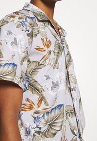 Camicia a maniche corte in tessuto grigio chiaro con un motivo floreale tropicale, con foglie verdi e blu e fiori arancioni e bianchi.