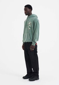 Groene hoodie met een voorzak en bloemenontwerp; gecombineerd met zwarte, wijde cargobroek en zwarte sneakers.