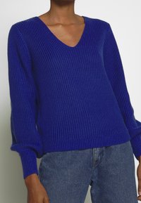 Blå stickad tröja med V-ringning, texturerat mönster, ribbade ärmslut och kant, som matchas med jeans.