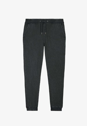 Femme aux cheveux blonds bouclés portant un t-shirt blanc, un pantalon de survêtement gris foncé avec cordon, et des baskets blanches, debout les mains dans les poches.