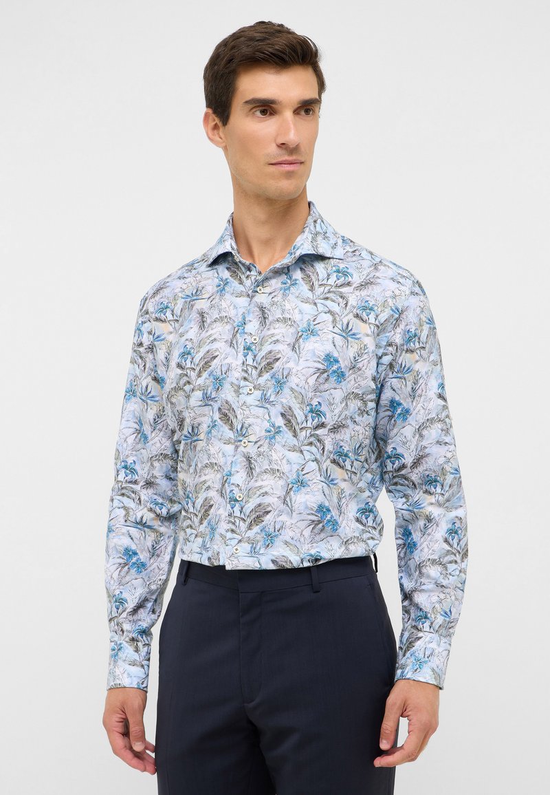 Man in een lange mouwen overhemd met blauwe en grijze bloemenprint, gestoken in donkere marineblauwe broek, staande tegen een witte achtergrond.