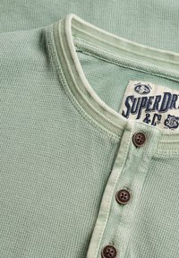 Camisa henley de punto waffle de color verde claro con tres botones marrones y una etiqueta visible de la marca Superdry dentro del cuello.
