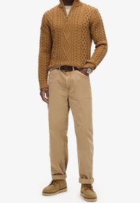 Gestrickter Pullover in gedämpftem Senfton mit hohem Kragen und Zopfmuster, kombiniert mit hellbeigen Chinos und hellbraunen Wildlederschuhen. Silberne Armbanduhr als Accessoire.