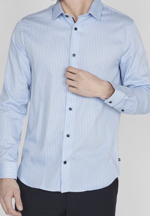 Homme portant une chemise à manches longues rayée bleu clair et blanc, ajustant le bouton du haut, sur un fond uni.