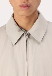 Light beige jacket with a large, flat collar, front zip closure, and contrasting inner lining texture. Smooth fabric with minimal detailing.