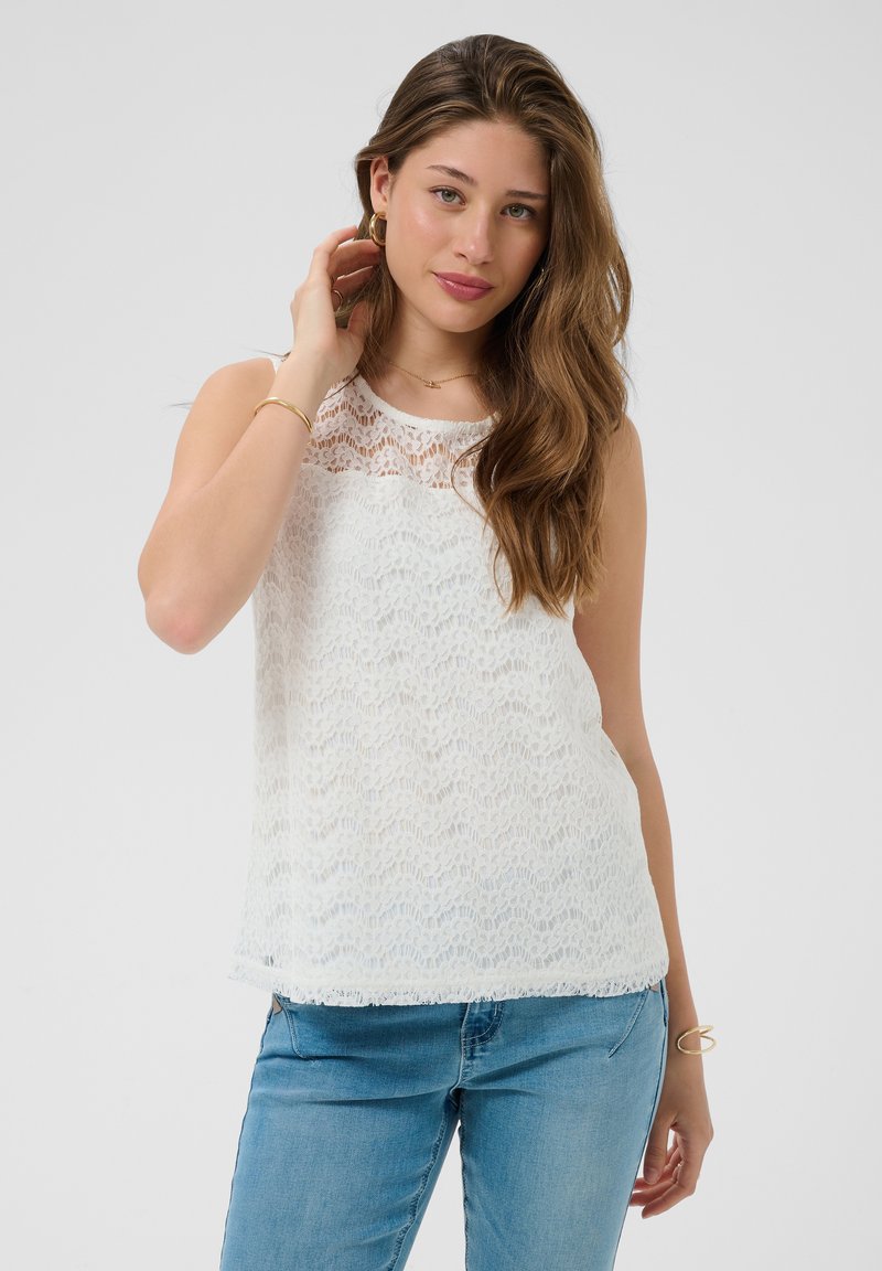 Jeune femme aux longs cheveux bruns, portant un top en dentelle blanche sans manches et un jean bleu, touchant ses cheveux sur un fond uni.