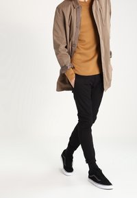 Brązowy długi płaszcz na musztardowym swetrze, czarne dopasowane spodnie i czarne tenisówki z białymi podeszwami. Widoczne skarpetki w kropki.