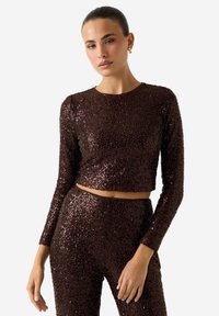 Top corto marrone in paillettes con maniche lunghe, caratterizzato da uno scollo rotondo e un design aderente. La superficie riflette la luce con una texture lucida.