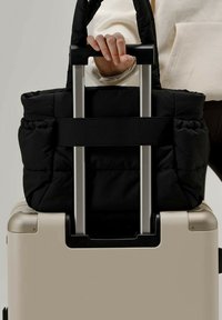 Musta tikattu kantokassi, jossa on kaksinkertaiset kahvat, kiinnitetty vaaleanbeigeen matkalaukkuun hihnalla, joka lepäilee tasaisella pinnalla. Hopeinen vetokahva näkyvissä.