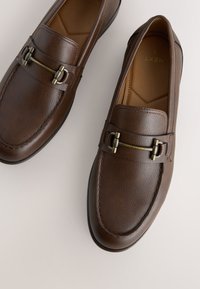 Paire de mocassins en cuir marron avec détail en métal et coutures sur une surface claire.