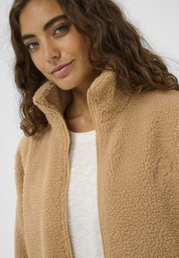 Kollane fliisjakken kõrge kraega ja tõmbluku sulgemisega. Omab pehmet, tekstuurset pinda ning peenet tikandit. Allpool on valge särk.