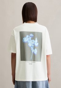 T-shirt en coton blanc avec un grand imprimé graphique d'une fleur dans des tons de bleu, avec du texte en dessous sur un fond gris. Style ample.