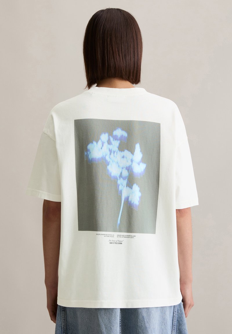 T-shirt en coton blanc avec un grand imprimé graphique d'une fleur dans des tons de bleu, avec du texte en dessous sur un fond gris. Style ample.