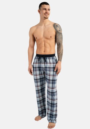 Oberkörperfreier Mann mit tribalem Tattoo am linken Arm, trägt blaue und weiße karierten Schlafanzughosen, steht barfuß und schaut mit einem leichten Lächeln zur Seite.