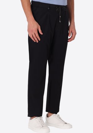 Uomo con pantaloni neri con coulisse e sneakers bianche, in piedi con le mani rilassate lungo i fianchi su sfondo uniforme.