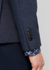 Närbild av en persons arm som bär en texturerad marinblå kavaj med fyra svarta knappar på ärmen och en skjortärms med blommönster som sticker ut.