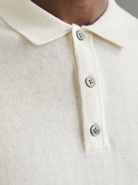 Polo-shirt crème clair avec une texture tricotée douce, un col classique et trois boutons de couleur argent.
