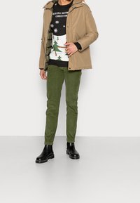 Giacca puffer beige con cappuccio, pantaloni in velluto a coste verdi, maglione natalizio nero con grafica che presenta alberi, e stivali neri alla caviglia con suola spessa.