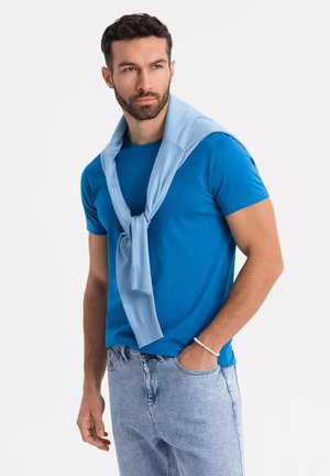 Blaues Rundhals-T-Shirt, kombiniert mit leicht blauem Pullover, der über die Schultern gelegt ist, getragen mit hellfarbigen Jeans. Sanfte Stofftextur.
