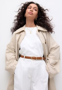 Beige trench coat, white top, brown leather belt with gold buckle, high-waisted white trousers, gold pendant necklace, wavy dark hair.