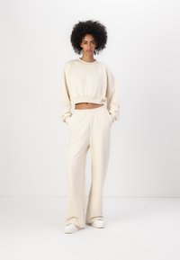 Beige cropped genser med lange ermer og elastisk kant, kombinert med beige vide bukser med et brodert logo. Hvite sko.