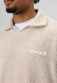 Beige fleece pullover met een gestructureerd oppervlak, voorzien van een ondiepe kraag en een wit geborduurd logo aan de linkerkant van de borst.