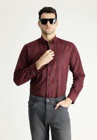 Camisa de botón roja con un estampado de patrones y sutiles rayas verticales, combinada con jeans de mezclilla grises. El modelo lleva gafas de sol negras.