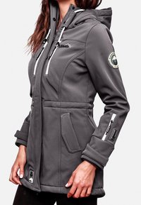 Veste softshell grise avec capuche, accents de fermeture éclair blancs, poches latérales et écusson logo sur la manche. Présente une coupe ajustée avec un tissu texturé.