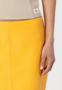 Geel leren rok met een gladde textuur en een zijnaad. Boven is een lichtbeige geribde top zichtbaar met een klein logo-label.