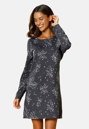 Lächelnde Frau in einem langärmeligen dunkelgrauen Kleid mit kleinen weißen und pinkfarbenen Blumenmuster, die mit einer Hand ihr Haar berührt.