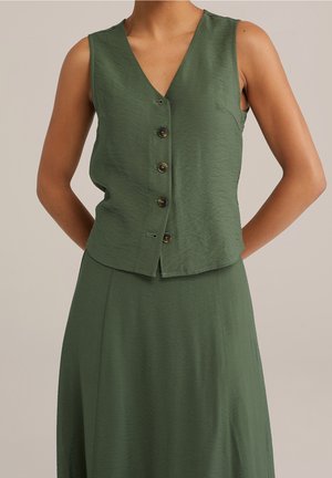 Veste sans manches vert olive boutonnée associée à une jupe taille haute assortie portée par une personne avec les mains dans le dos.