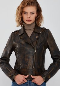 Giacca da moto in pelle marrone con una texture vissuta, cerniera asimmetrica, bottoni a pressione e tasche con zip, abbinata a un dolcevita.