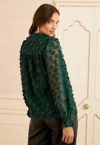 Zelená halenka s puntíky, vrstvou texturované látky, průhlednými rukávy a nařasenými manžetami, v kombinaci s černými kalhotami.