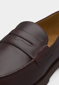 Σκούρο καφέ δερμάτινο παπούτσι τύπου penny loafer με λεπτομερή ραφές και σχέδιο moc toe, εμφανιζόμενο από λοξή κοντινή οπτική γωνία.