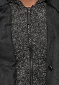 Zwarte zip-up jas met een gestructureerde grijze binnenlaag; beschikt over een contrasterende zwarte rits en knoopaccent, met een aangehechte capuchon.