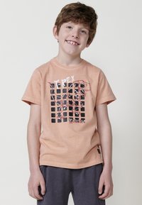 Persikų medvilnės marškinėliai su T-Rex grafika, papuošta juodais ir raudonais akcentais, o fone matomas tinklo raštas. Marškiniai su apvalia iškirpte ir trumpomis rankovėmis.