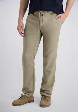Mann trägt beige Kordelhose und beige Freizeitschuhe, steht mit einer Hand in der Tasche vor schlichtem Hintergrund.