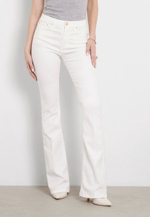 Woman wearing high-waisted white flare jeans and beige pointed-toe heels, standing against a plain light background.