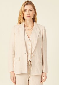 Blazer de lino en tono beige claro con un diseño entallado, solapas muescas y dos bolsillos frontales. Llevado sobre un chaleco a juego con cierre de botón.
