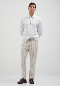 Homme debout portant une chemise blanche à manches longues avec des détails de boutons, un pantalon rayé beige et noir, et des chaussures brun foncé.