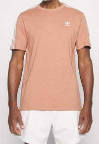 Ljus persikofärgad bomulls-t-shirt med rund halsringning, korta ärmar och vit Adidas-logga på bröstet. Har tre vertikala vita ränder på axlarna.