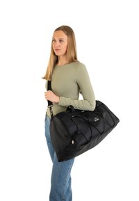 Black duffel bag with textured surface, dual handles, and adjustable shoulder strap. Features elastic cord detail on the front.